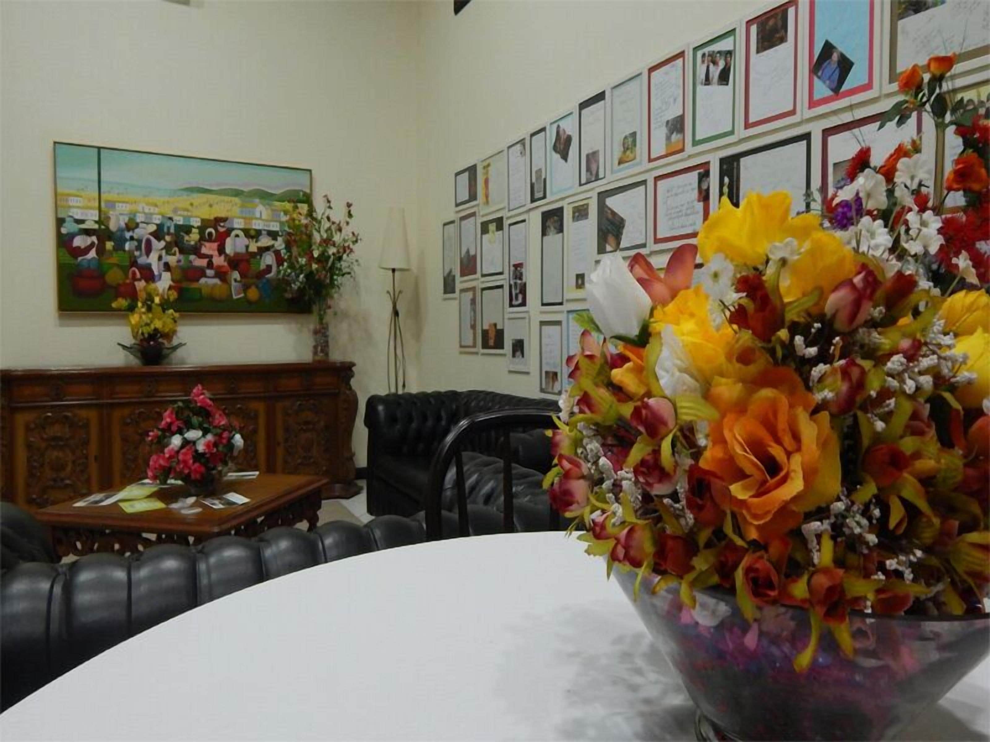 Uma sala de conferências com um arranjo floral grande sobre uma mesa, um sofá de couro preto, um armário de madeira com um vaso de flores, e uma parede com fotos emolduradas e uma pintura.