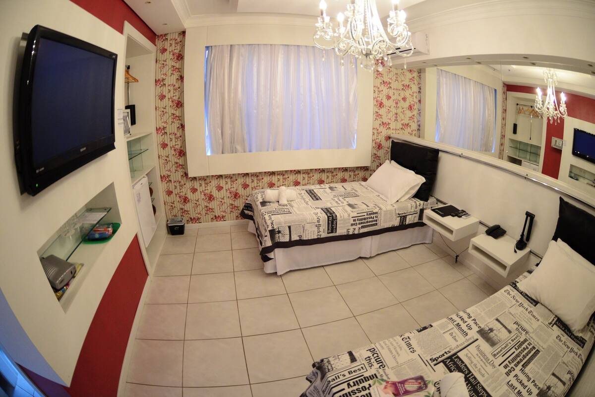 Un dormitorio con cama, televisor, mesita de noche y espejo, además de un ventanal con cortinas.