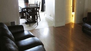 A living room with a dark leather sofa, a dining table with chairs, and a wooden floor. - Bk Guest house (Anacortes)