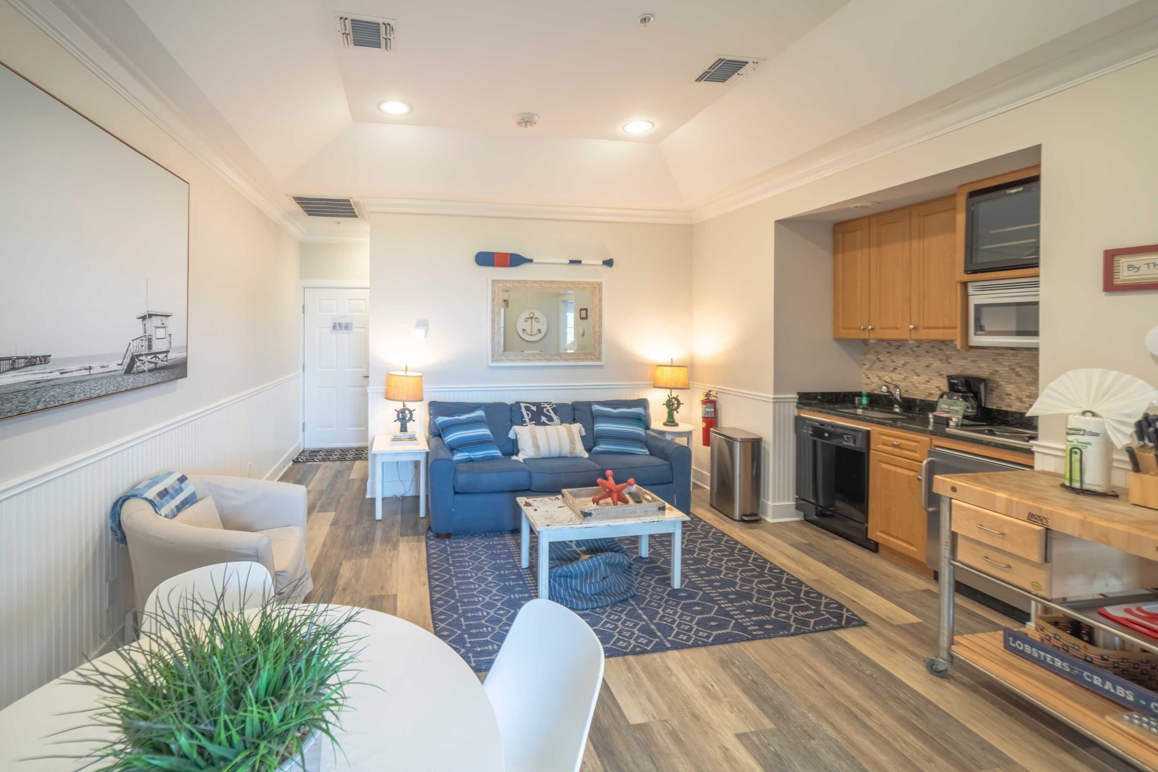 A living room with a sofa, coffee table, and a dining area with a table and chairs. There is a kitchen with appliances and cabinets.