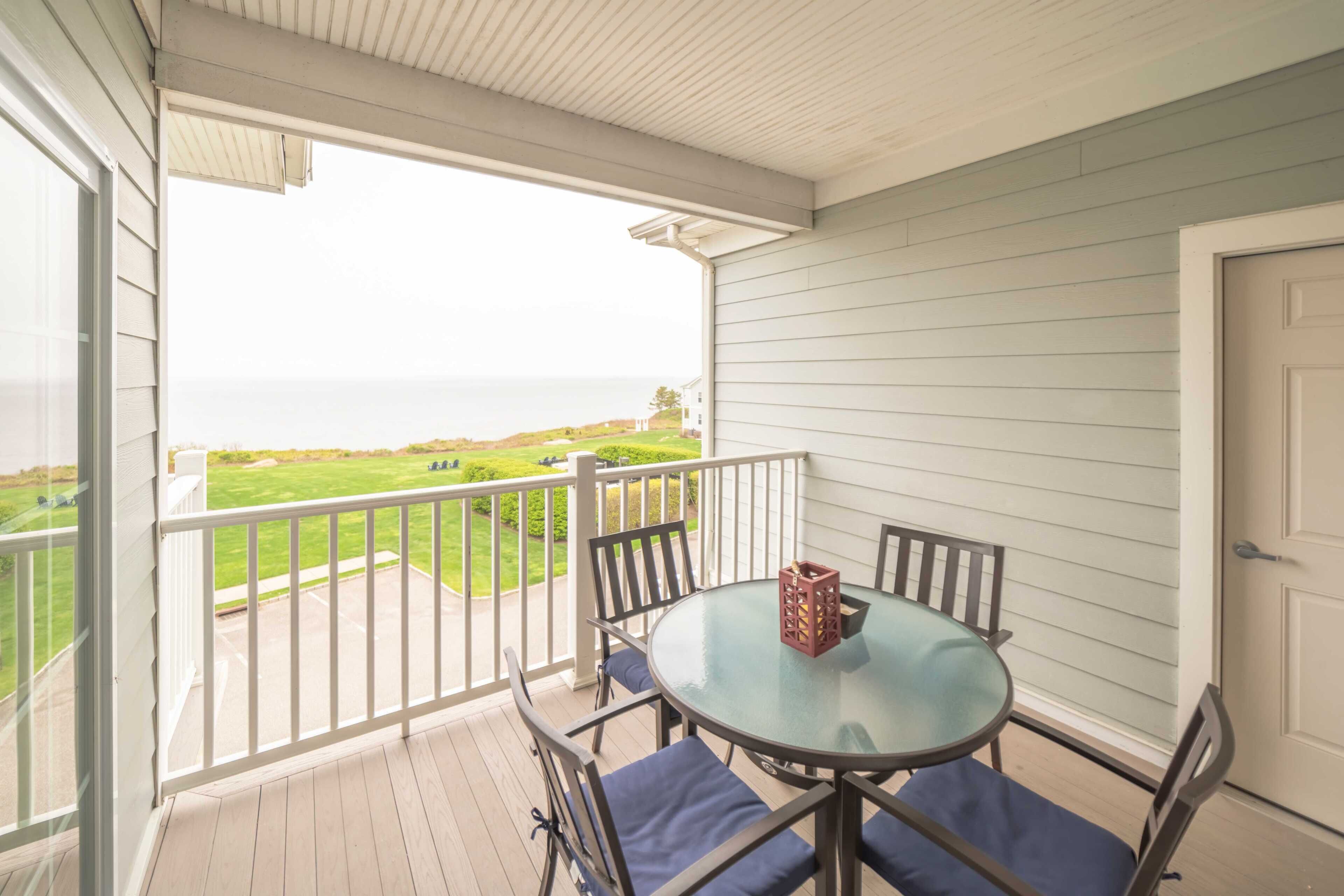 A covered balcony with a glass table and chairs, overlooking a green landscape and the sea.