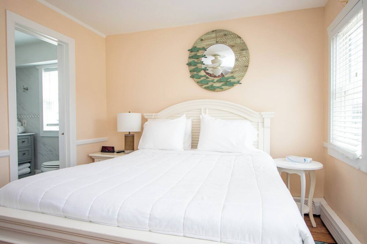 A bedroom with a bed, a bedside table with a lamp, a round mirror on the wall, and a window with blinds.