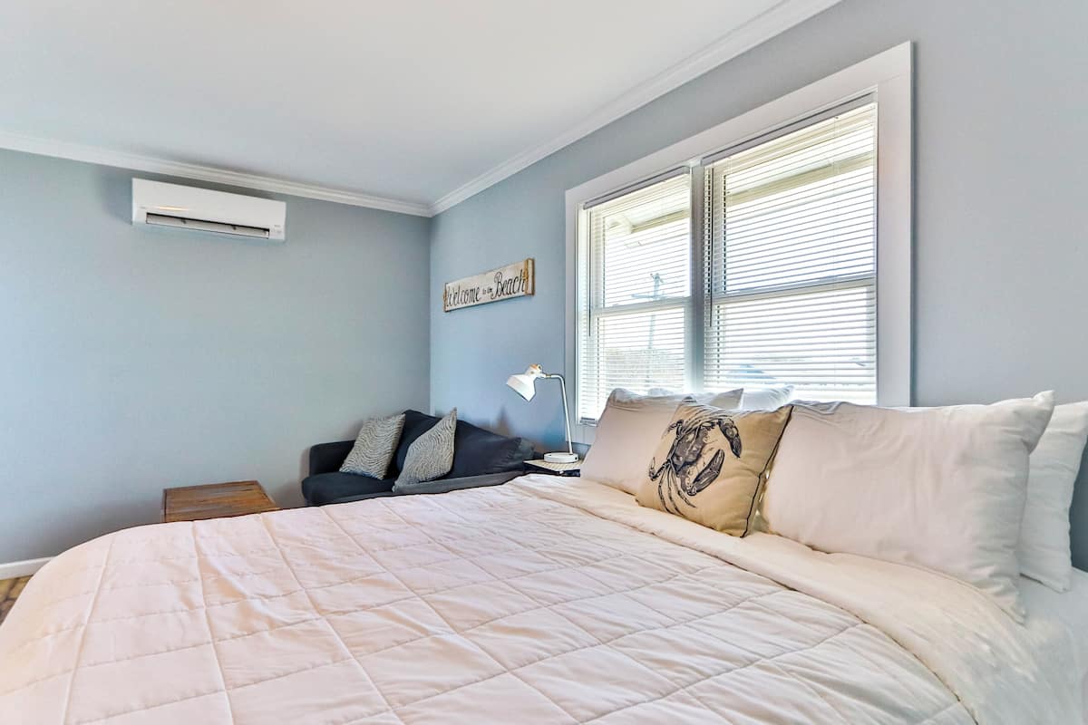 A bedroom with a bed, a nightstand, a lamp, a window with blinds, and a wall-mounted air conditioner.