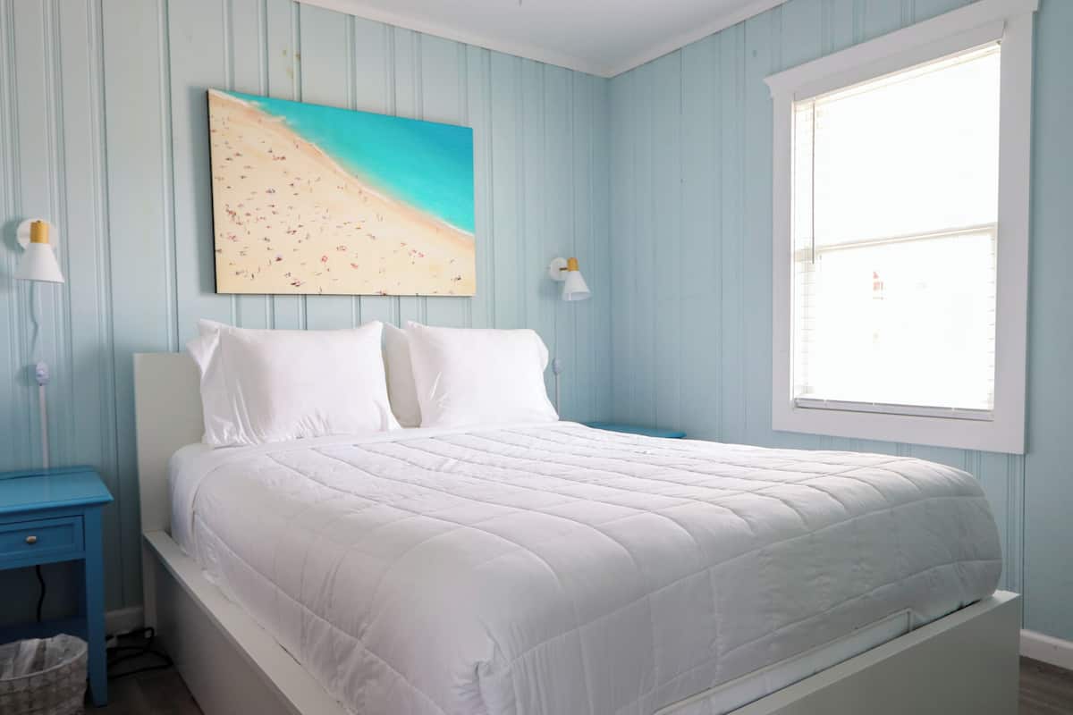 A bedroom with a bed, a painting of a beach, a window, and a nightstand.