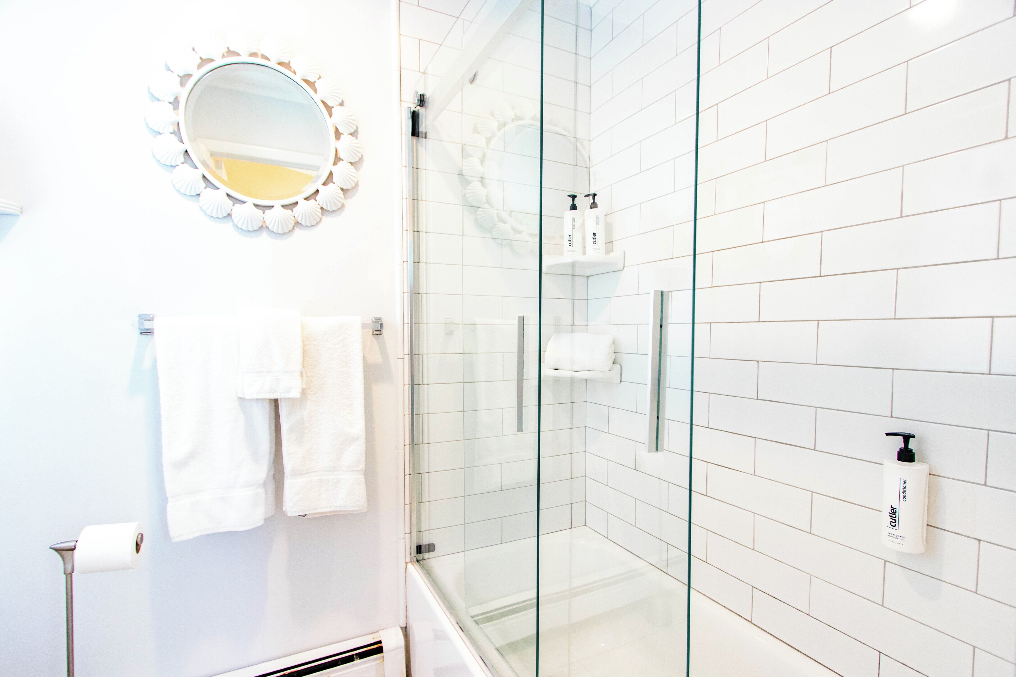 Un baño moderno con azulejos blancos, cabina de ducha de vidrio, espejo redondo con marco ondulado y un perchero para toallas.