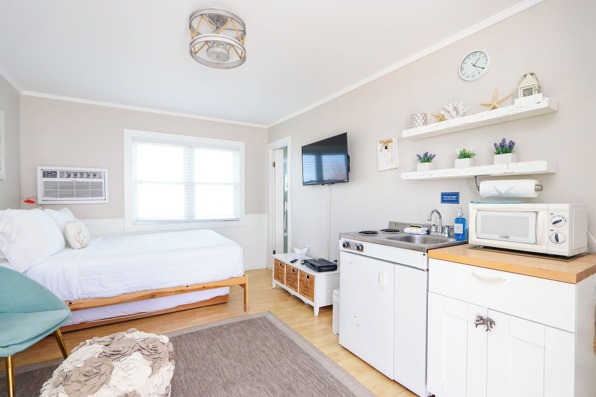 A bedroom with a bed, bedside table, and a TV. There is a kitchen with a microwave, sink, and cabinets.