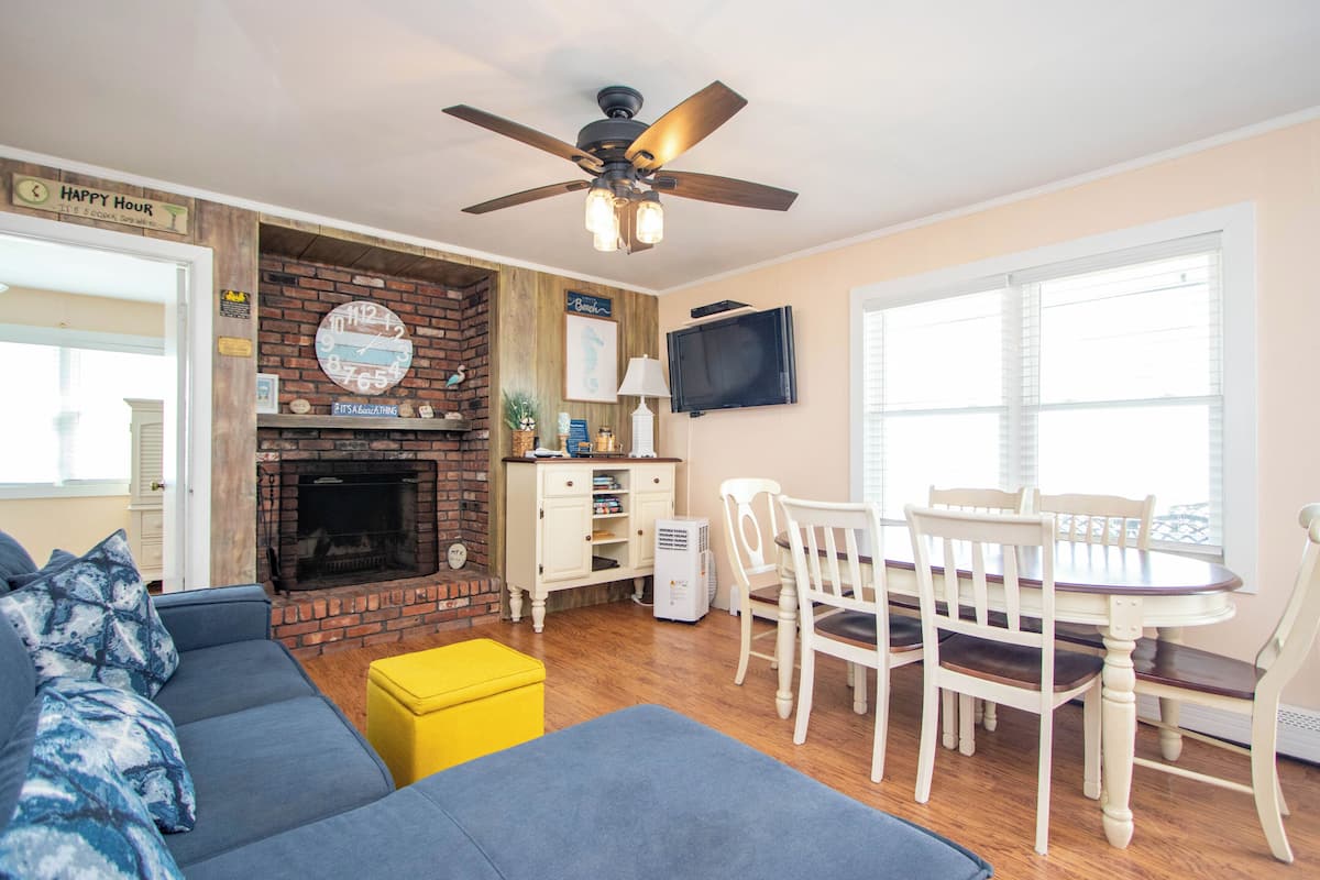 A living room with a fireplace, a ceiling fan, a dining table with chairs, and a TV mounted on the wall.