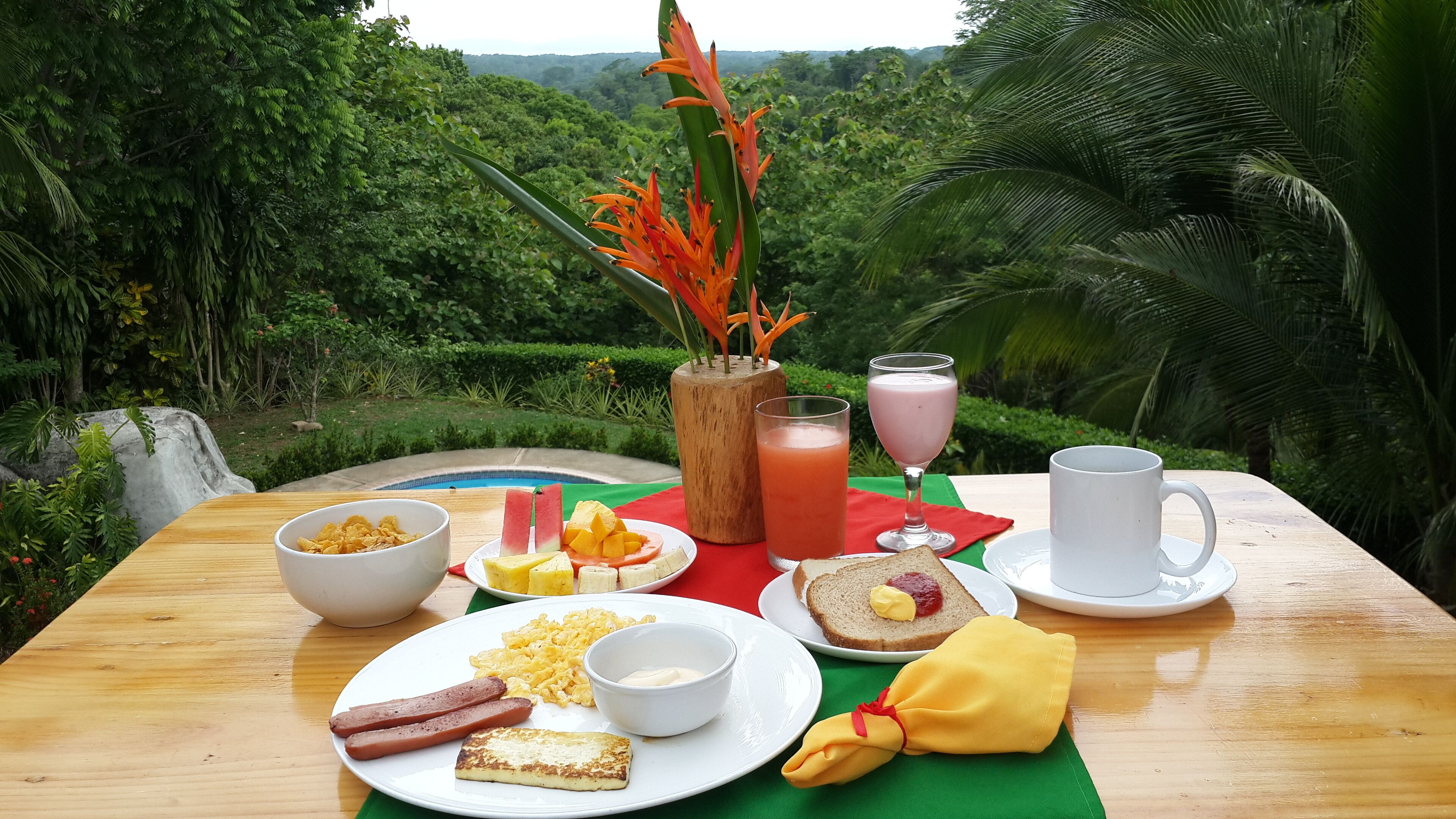 Una mesa de desayuno con diversas comidas y una bebida, frente a un paisaje verde exuberante.