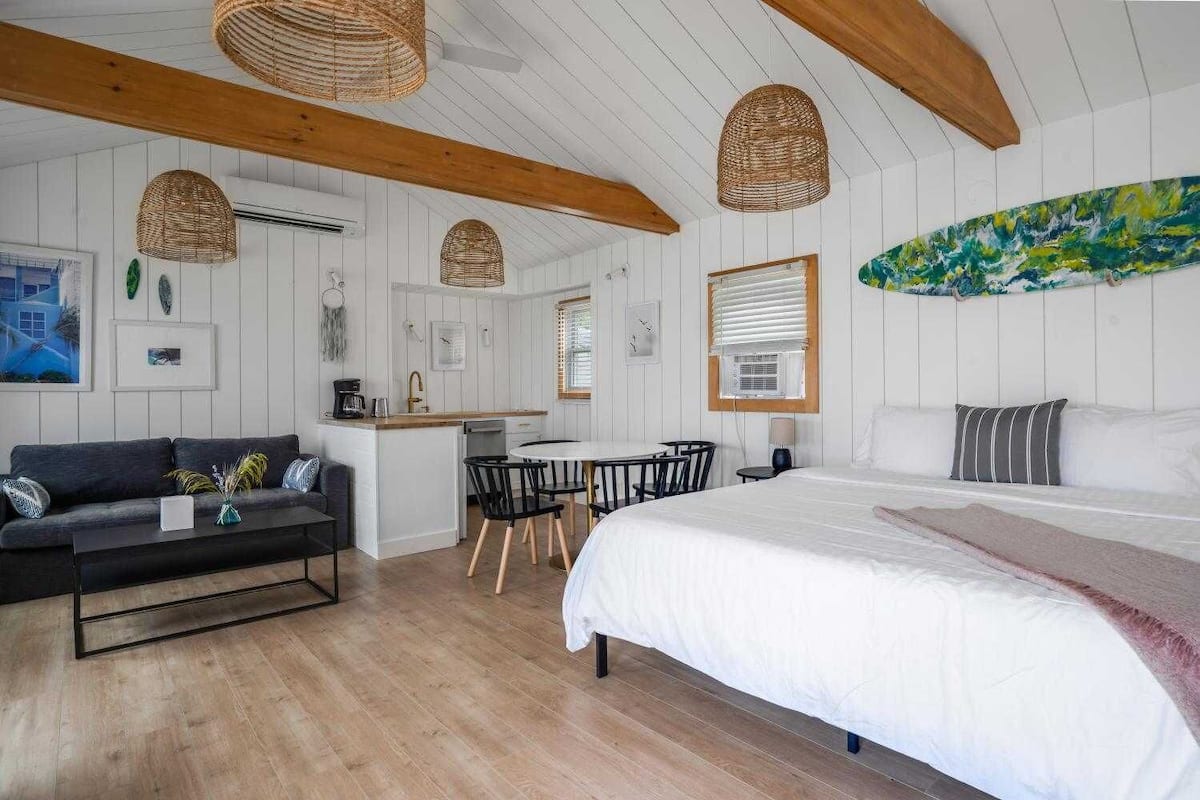 Ein Zimmer mit einem Bett, einem Schlafsofa, einem Esstisch mit Stühlen, einer Küchenzeile und einem großen Fenster mit Jalousien.