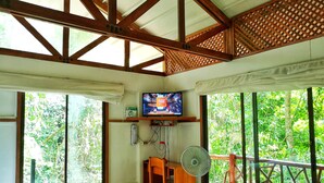 Ein Zimmer mit einem Bett, einem Schreibtisch, einem Fernseher und einem Ventilator. Es gibt einen Balkon mit Holzgeländer und Bepflanzung davor.