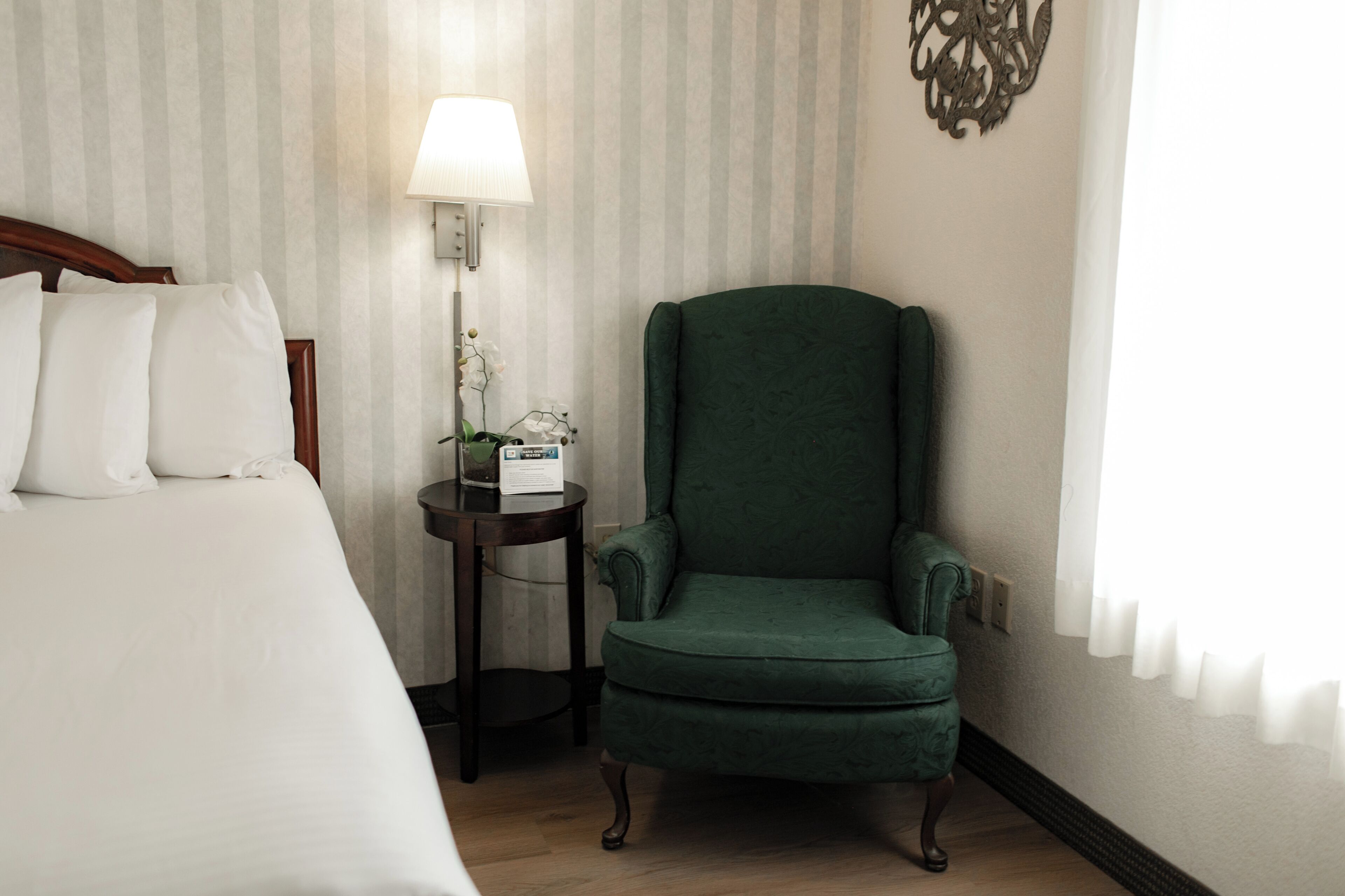 Une chambre d’hôtel avec un lit, un fauteuil vert, une table de chevet avec une lampe et un élément décoratif mural.