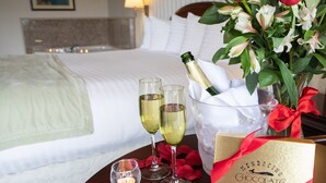 A hotel room with a neatly made bed, a bedside table with champagne, glasses, a candle, rose petals, and a chocolate gift box. - Emerald Dolphin Inn & Mini Golf (Fort Bragg)