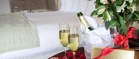 A hotel room with a neatly made bed, a bedside table with champagne, glasses, a candle, rose petals, and a chocolate gift box.