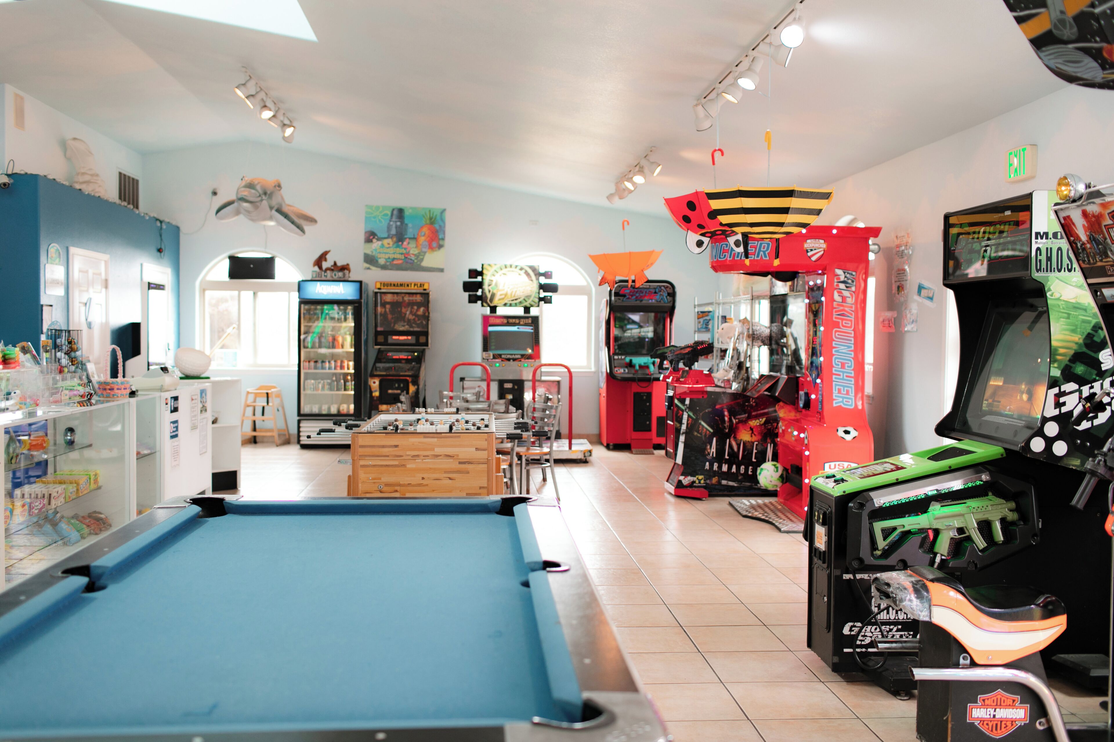 Mesa de billar, juegos de arcade y una máquina expendedora en una sala luminosa con techo alto y diversas decoraciones.