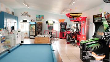 Mesa de billar, juegos de arcade y una máquina expendedora en una sala luminosa con techo alto y diversas decoraciones.
