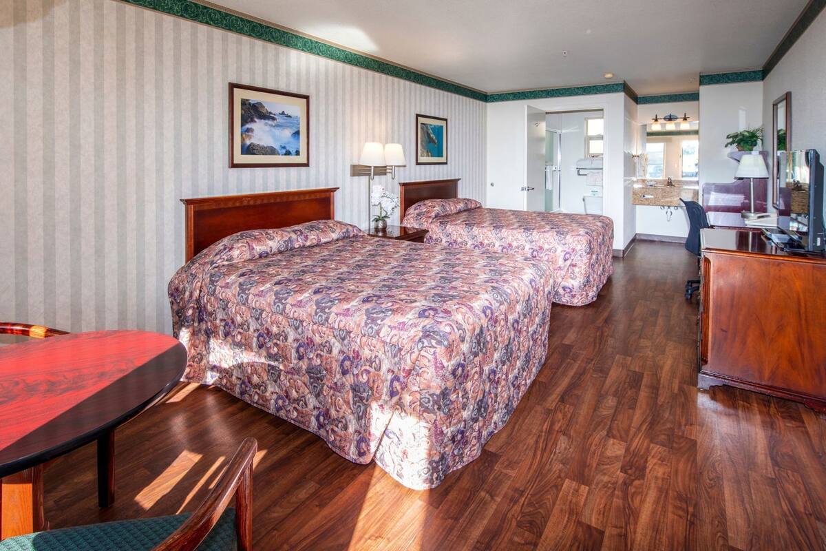 Quarto de hotel com duas camas, uma escrivaninha, uma cadeira, um armário e um banheiro visível por uma porta aberta.