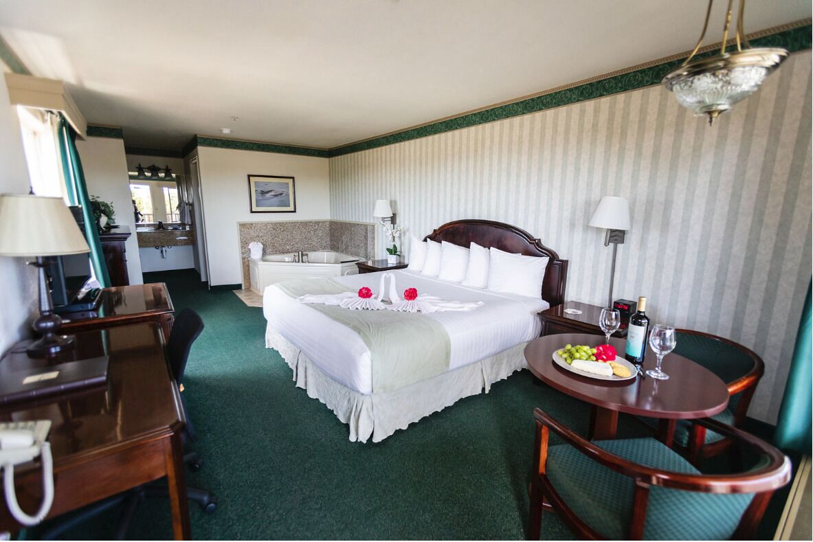 A hotel room with a bed, a desk, a chair, a small table with a plate of fruit and wine, and a mirror.