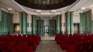 A large hall with rows of red chairs, green columns, and a central aisle leading to a door. - Phi Semesta (Semarang)