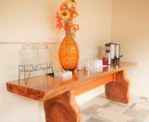Une table en bois, sur laquelle se trouvent un vase de fleurs, un distributeur d’eau et une machine à café.