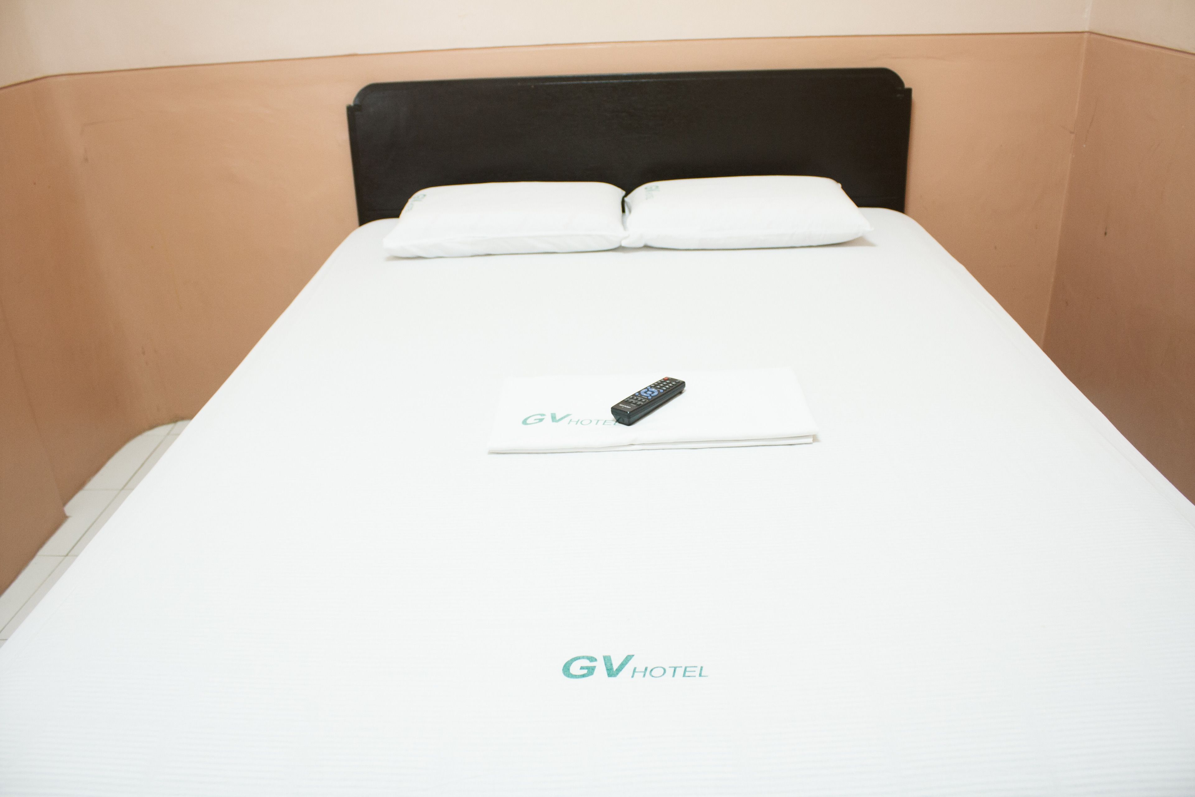 Een enkel bed met wit beddengoed en een afstandsbediening erop. Het bed heeft een zwart hoofdeinde en staat in een kamer met beige wanden.