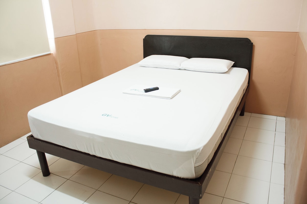 Una habitación pequeña con cama individual, cabecera negra, colchón blanco y un control remoto sobre la cama.