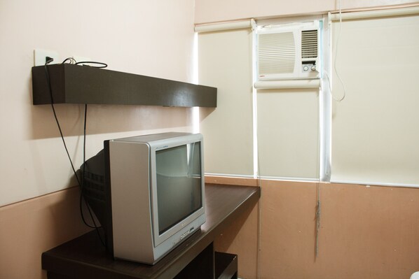 A television on a wooden stand in a room with beige walls and a window with blinds.