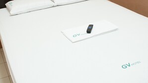 Een net opgemaakt bed met witte beddengoed en kussens, een afstandsbediening en het logo 'GV HOTEL' op de plaid.
