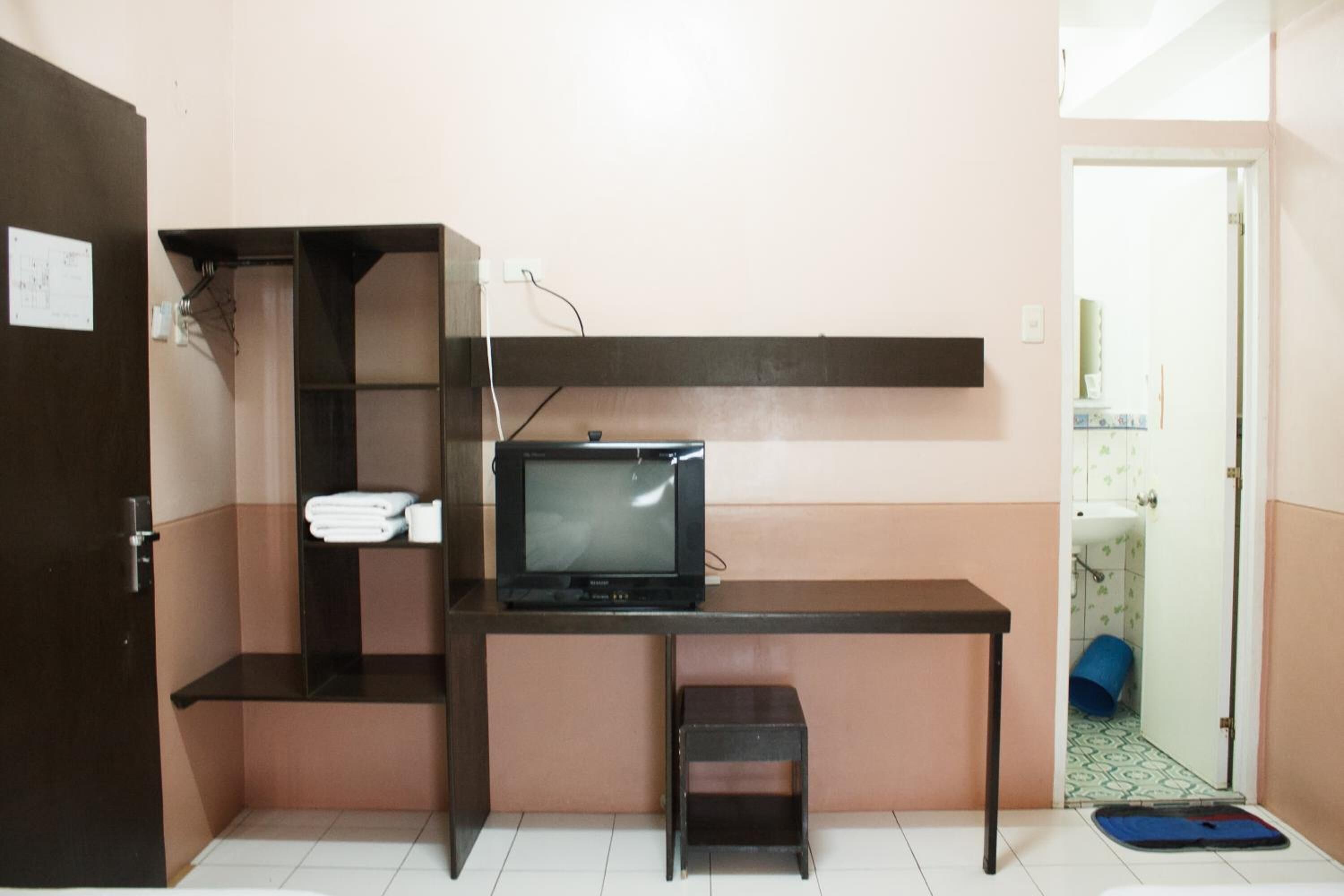Una habitación pequeña y funcional con una televisión sobre un escritorio, un taburete y un estante con toallas dobladas.
