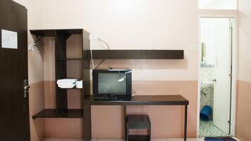 Una habitación pequeña y funcional con una televisión sobre un escritorio, un taburete y un estante con toallas dobladas.