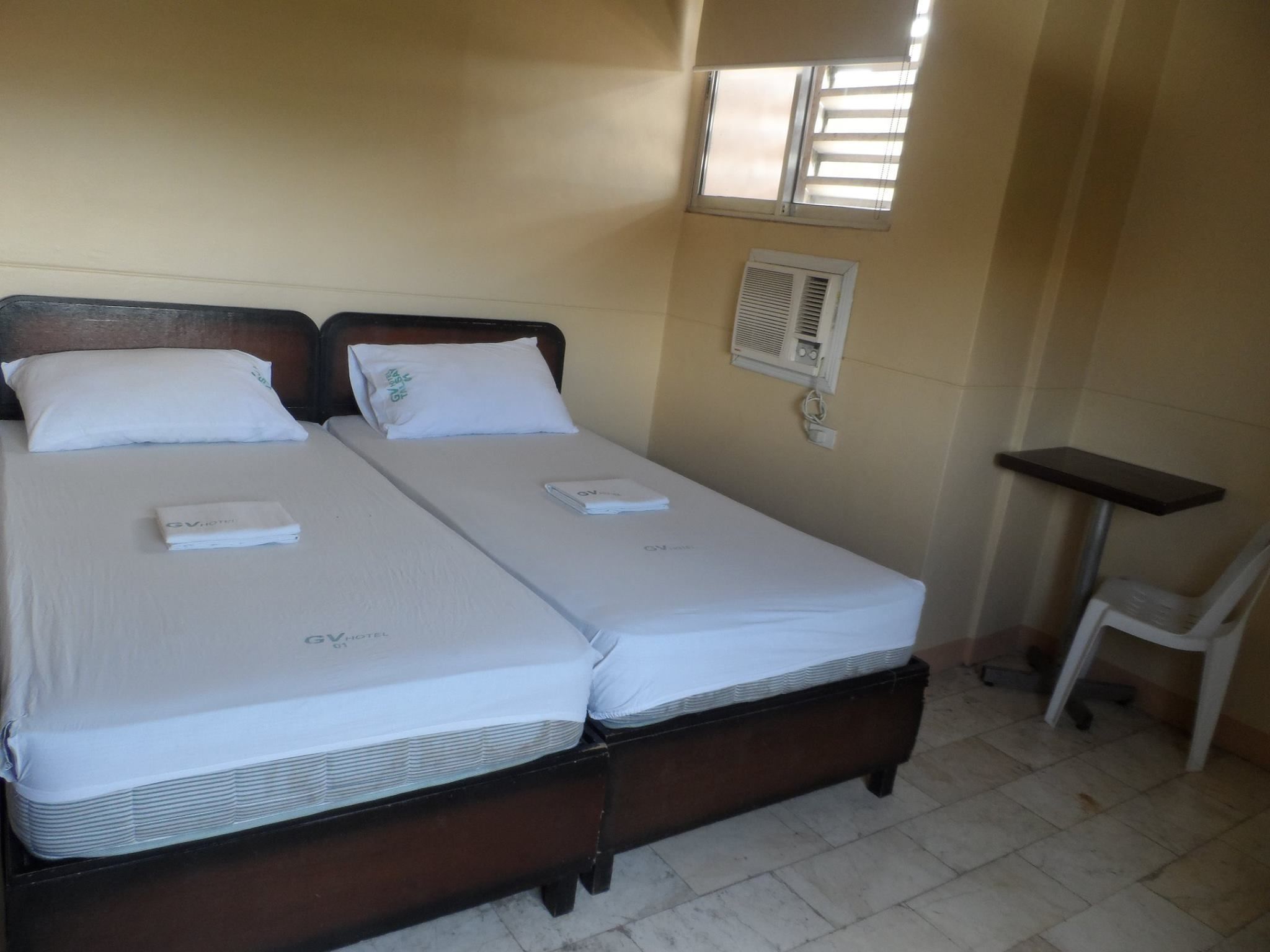 Un lit double avec du linge de lit blanc et une tête de lit en bois. Il y a une petite table de chevet avec une chaise, une fenêtre avec des stores et un climatiseur fixé au mur.