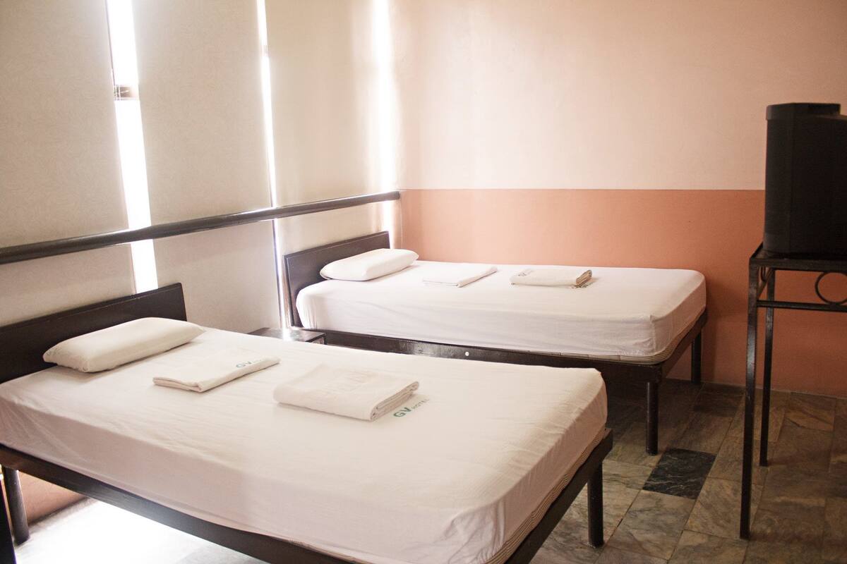 Two single beds with white linens and pillows, a television on a stand, and a tiled floor.
