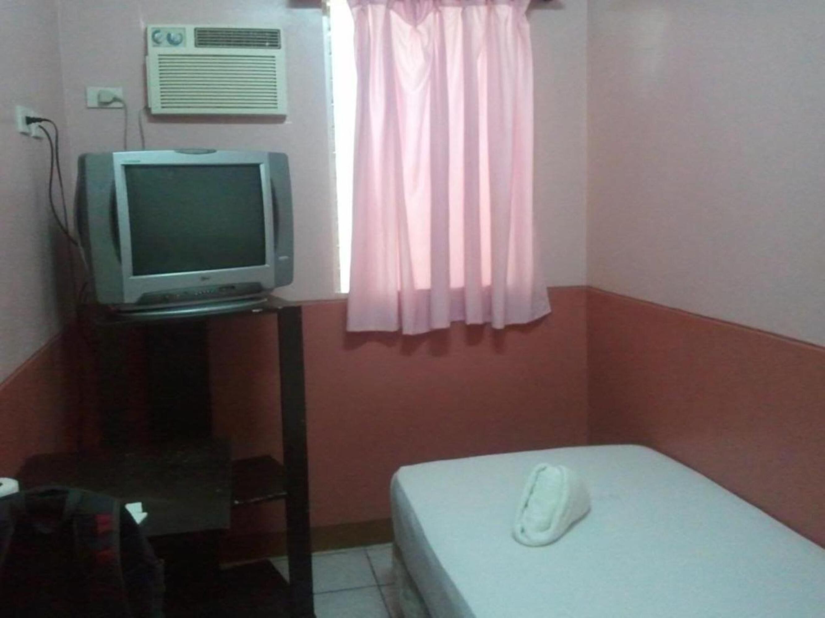 Quarto pequeno com uma cama, televisão em um suporte, janela com cortinas cor-de-rosa e ar-condicionado na parede.