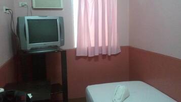 Quarto pequeno com uma cama, televisão em um suporte, janela com cortinas cor-de-rosa e ar-condicionado na parede.