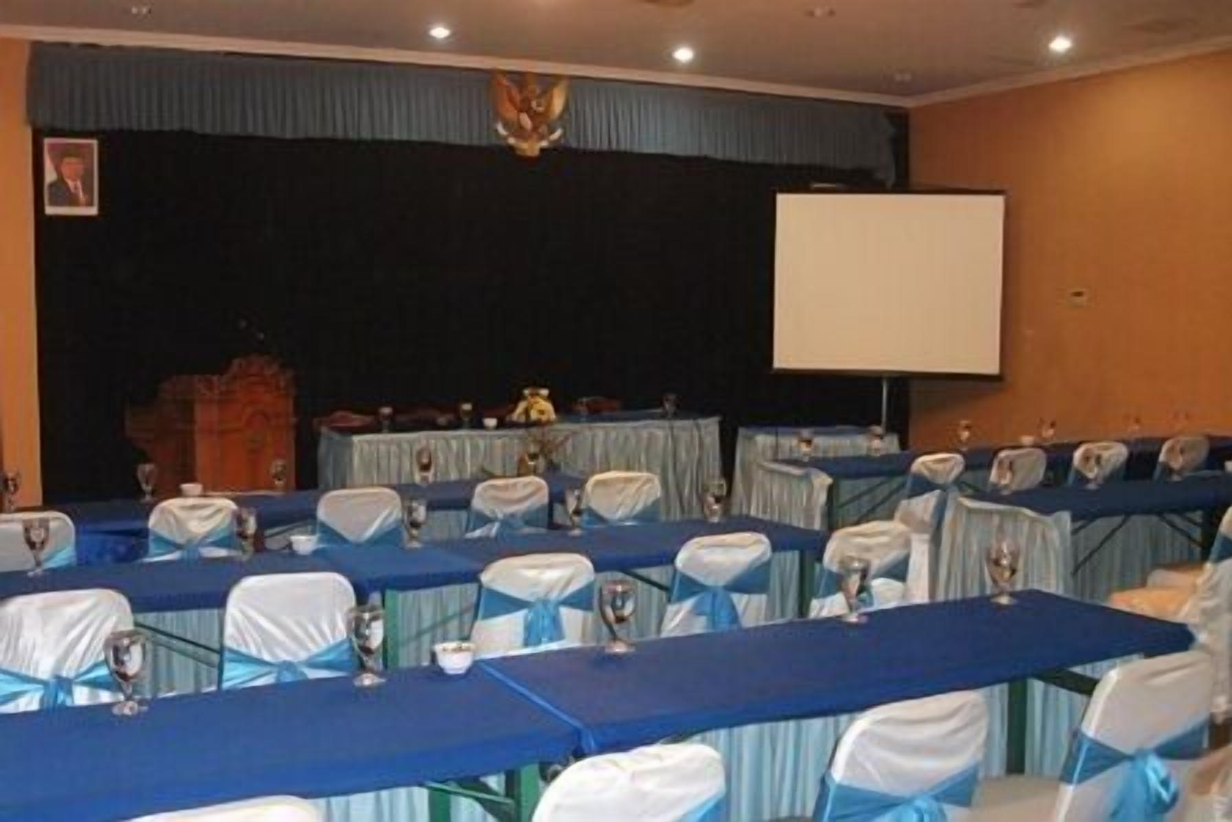 A conference room with rows of tables and chairs, a stage with a podium, and a screen.