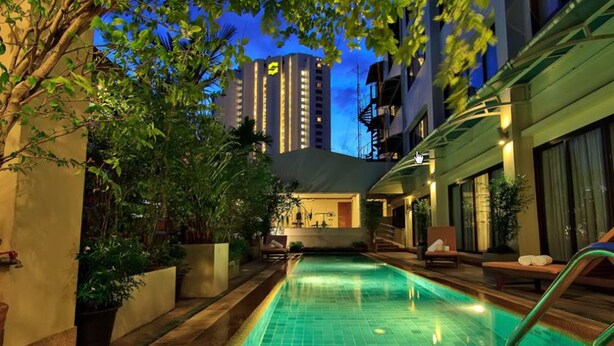 Una piscina al aire libre con sillones de descanso y plantas en macetas, rodeada de edificios y un cielo despejado.