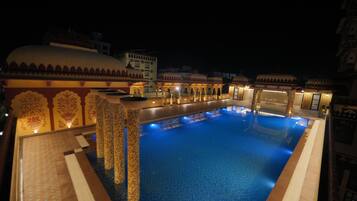 A swimming pool with a tiled deck and a fountain in the center, surrounded by ornate buildings with domed roofs.
