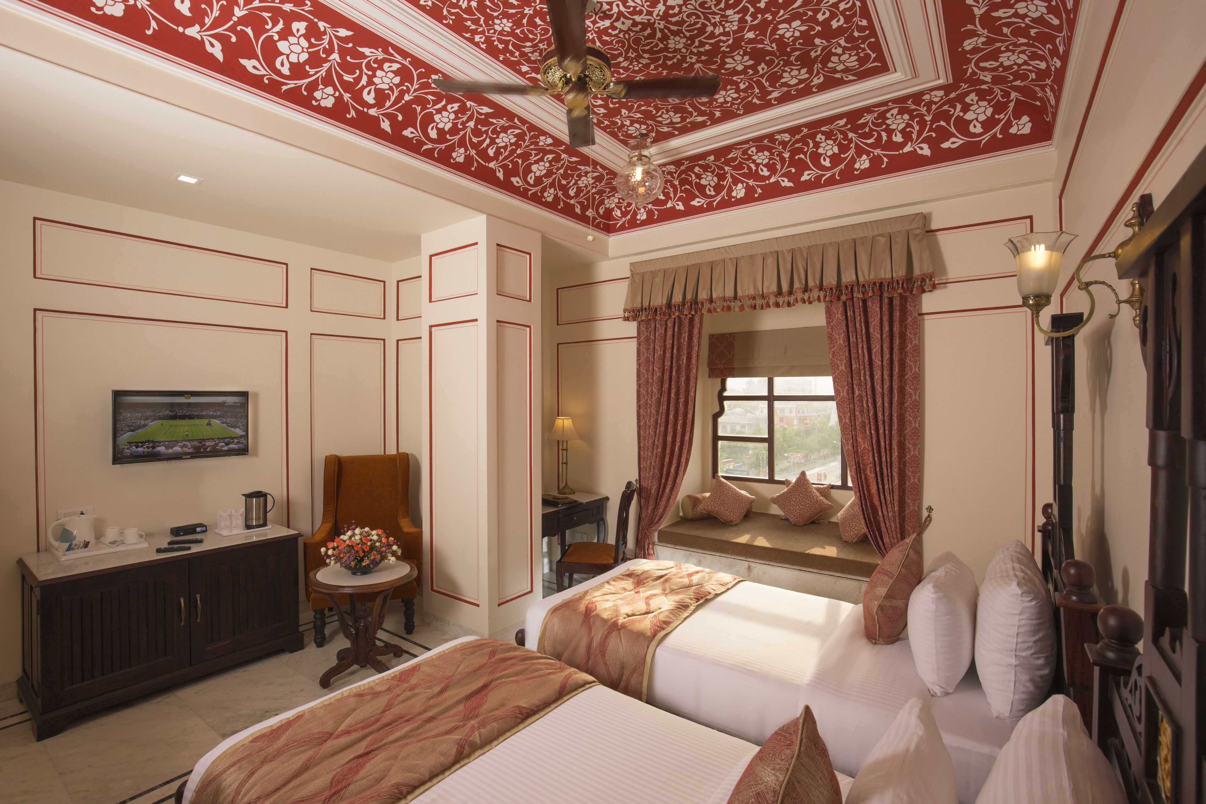 Hotelkamer met groot bed, bureau met lamp, stoel, raam met gordijnen en plafondventilator.