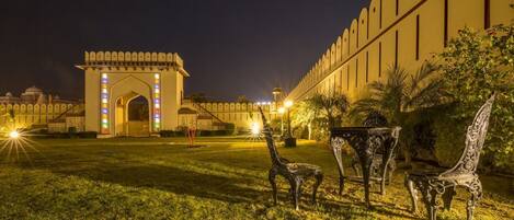 An outdoor area with a large, illuminated archway and a long, high wall. There are decorative chairs and a small table in the foreground.