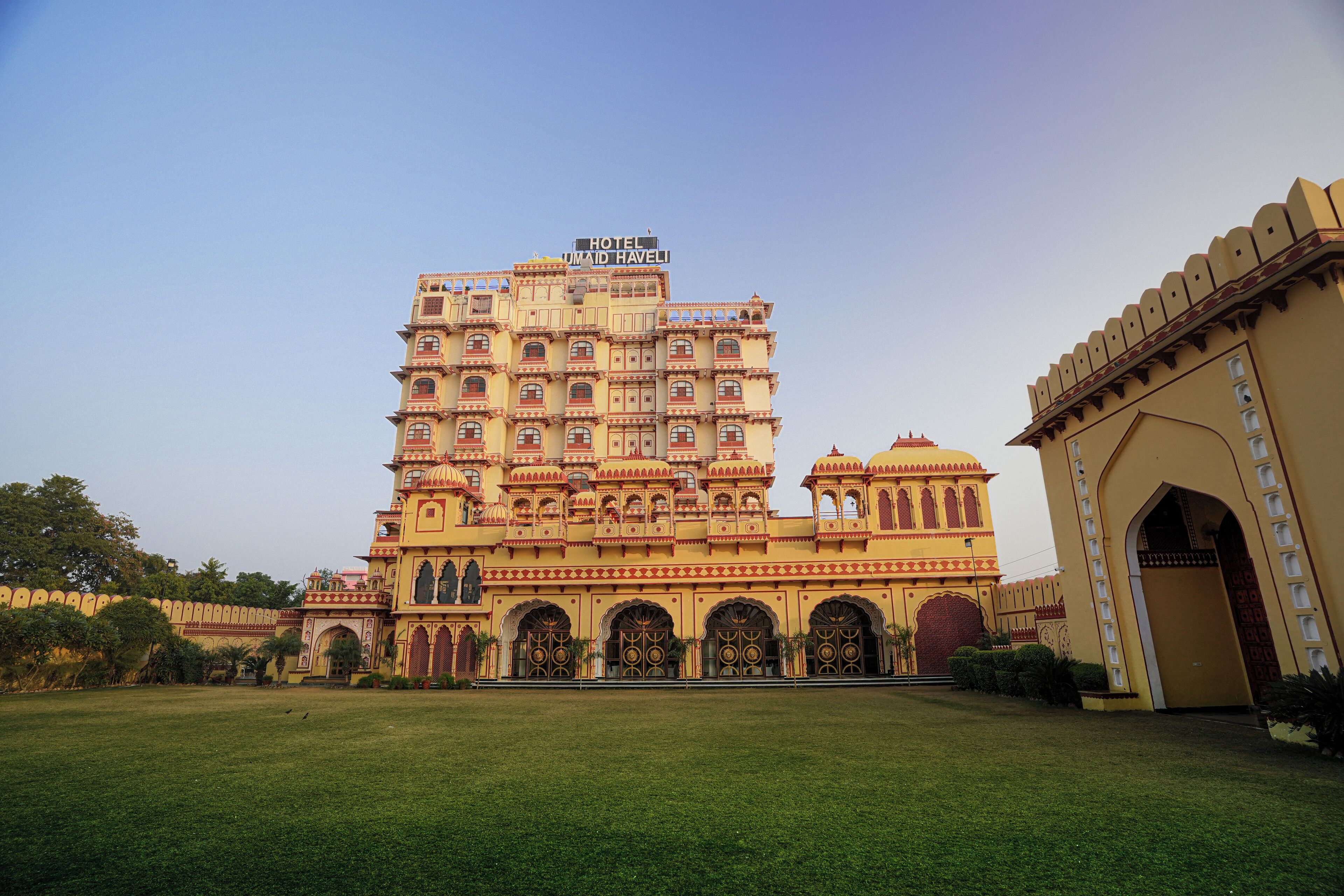 A large, multi-story building with a sign reading "Hotel Umaid Haveli." It features ornate architecture with multiple balconies and arched doorways.