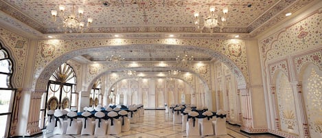 Un salón imponente con elaborados diseños florales en el techo, candelabros y filas de sillas blancas con cintas negras.