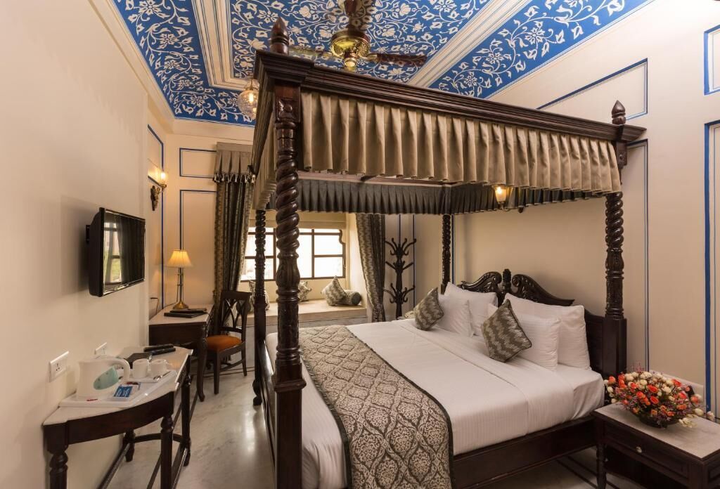 A four-poster bed with a canopy, a bedside table with a lamp, a desk with a chair, and a television on the wall.