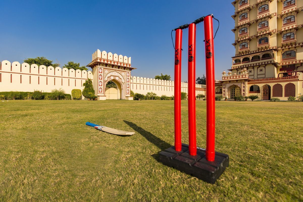 Un campo de críquet con tocones rojos y un bate azul frente a un edificio histórico con portales en arco y un muro alto.