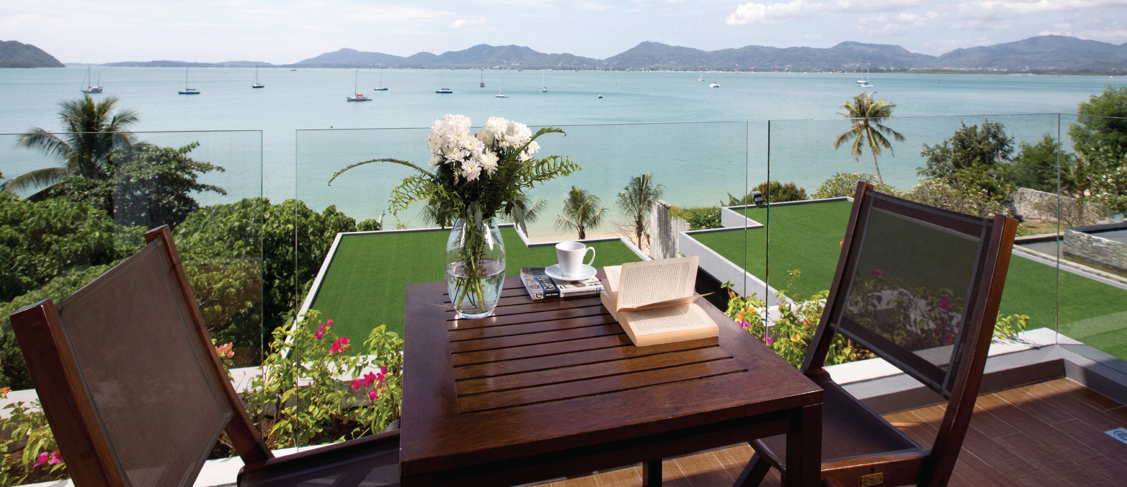 A wooden table with a vase of flowers, a cup, and a book on a balcony overlooking a body of water with boats.