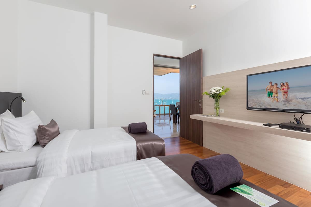 Een hotelkamer met twee bedden, een aan de muur bevestigde televisie en uitzicht op zee door een open deur.