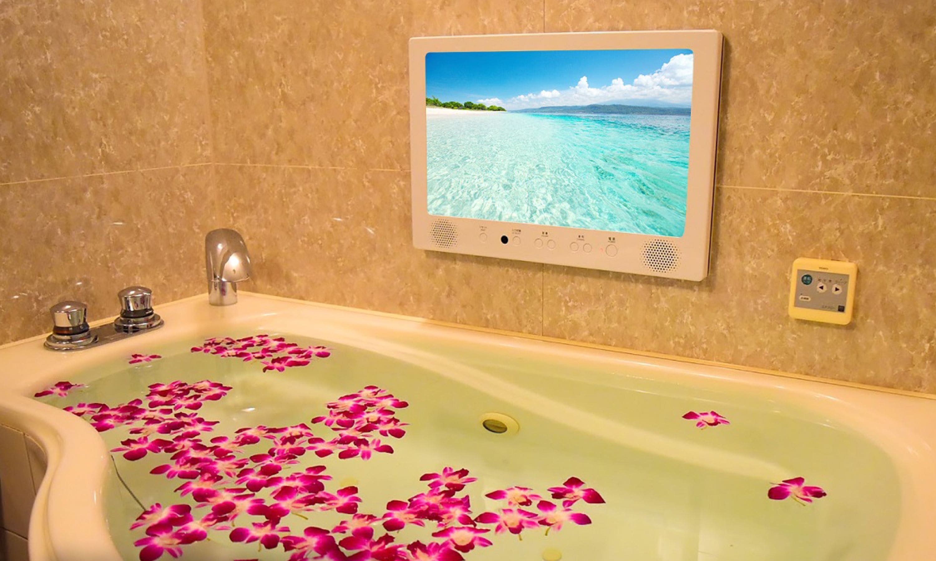 A bathtub with rose petals floating on the water, a wall-mounted TV displaying a beach scene, and a bathroom with beige tiles.