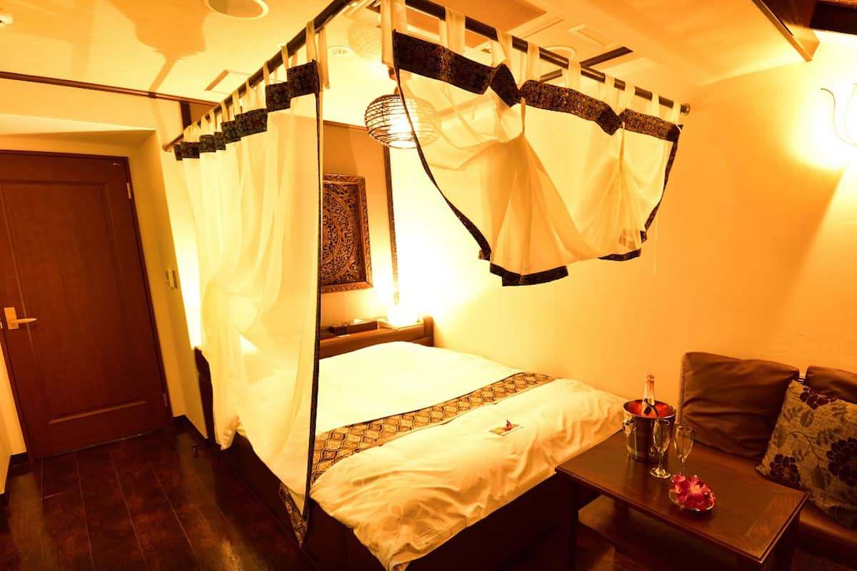 Una habitación con cama con dosel, una mesita de noche con una botella y vasos, y un sofá con un cojín.