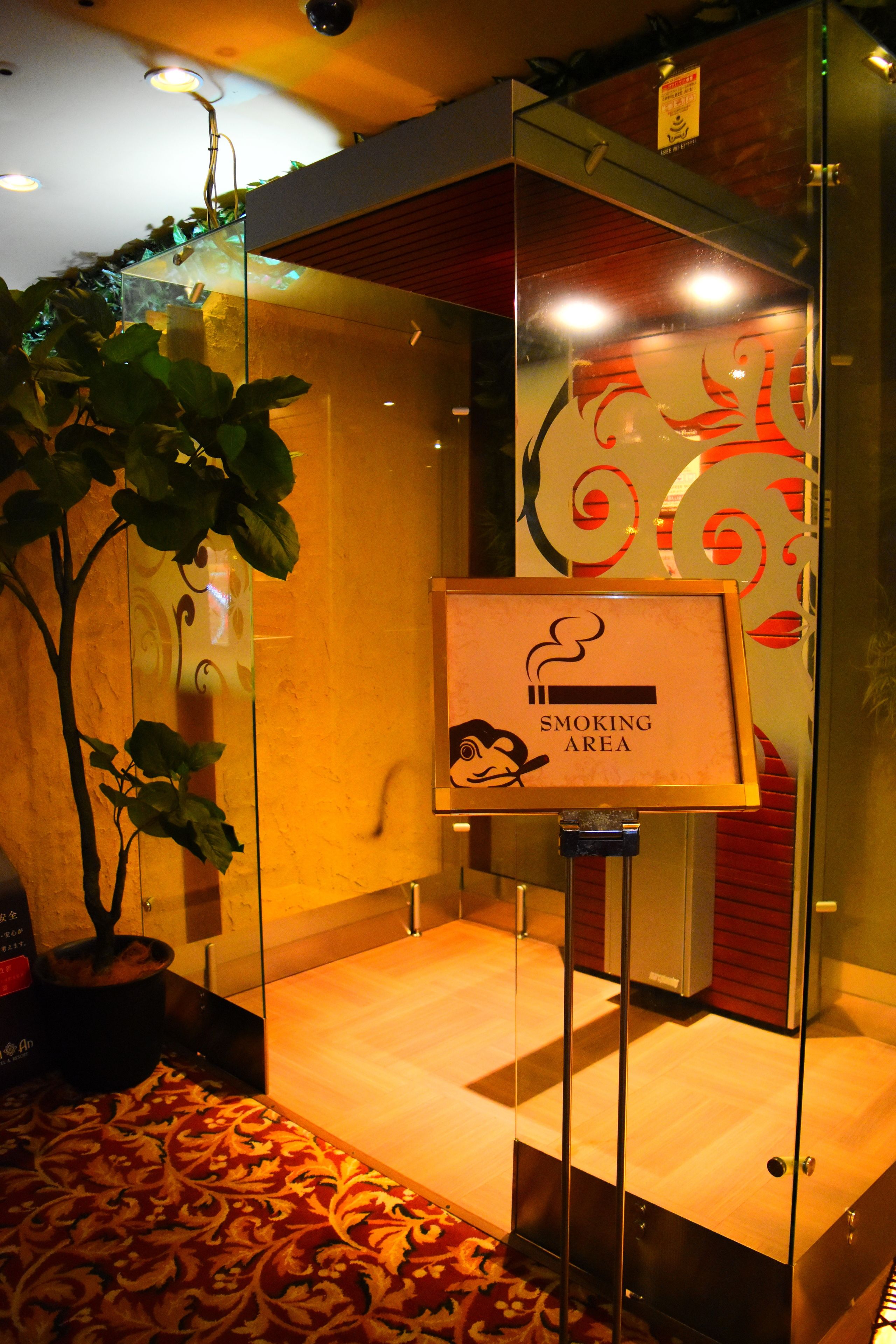 A smoking area with a glass partition, a potted plant, and a patterned carpet.