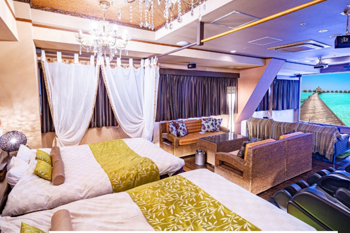 A luxurious room with a bed, sofa, massage chairs, and a large screen displaying a beach view.