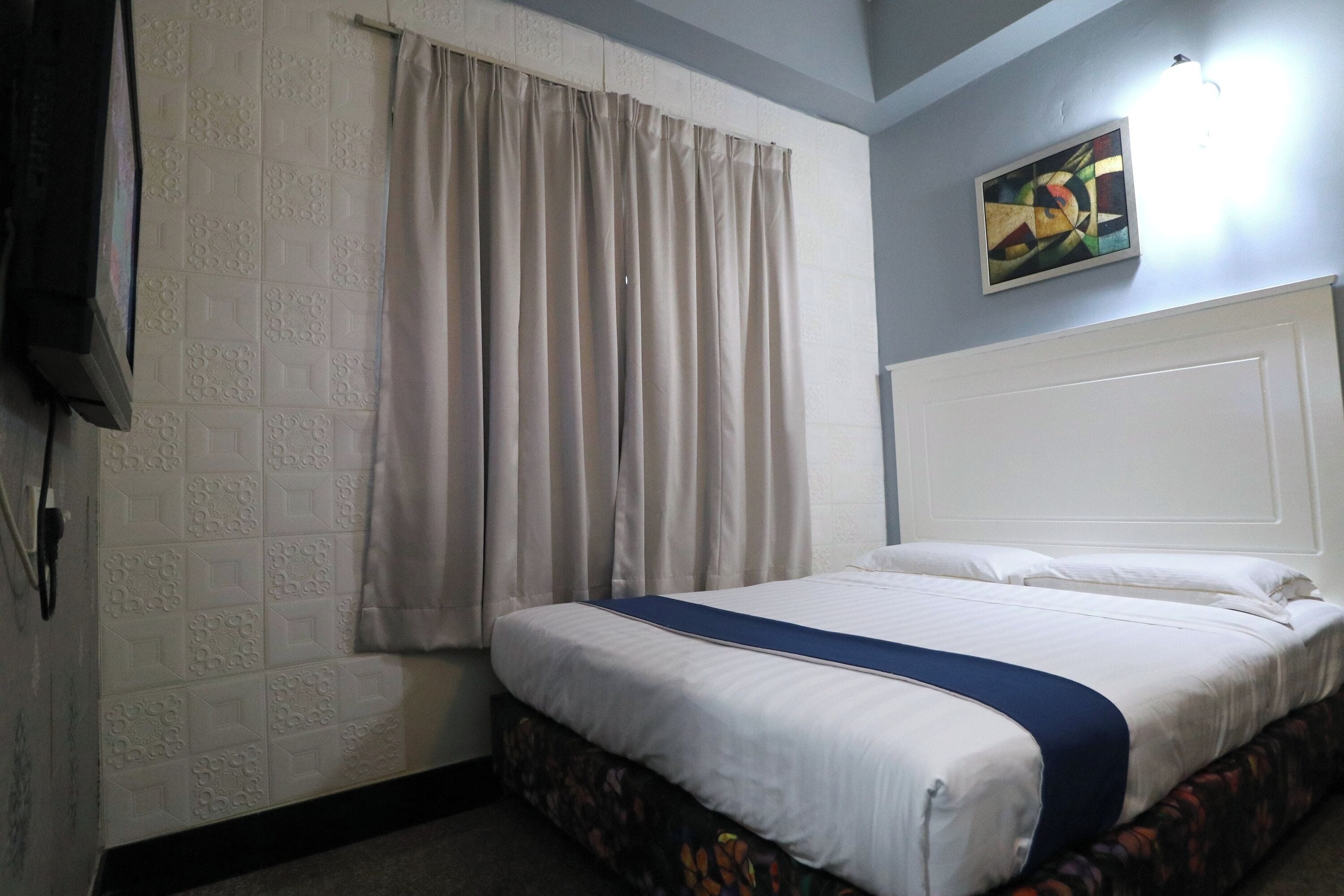 A hotel room with a bed, curtains, a painting on the wall, and a light fixture.
