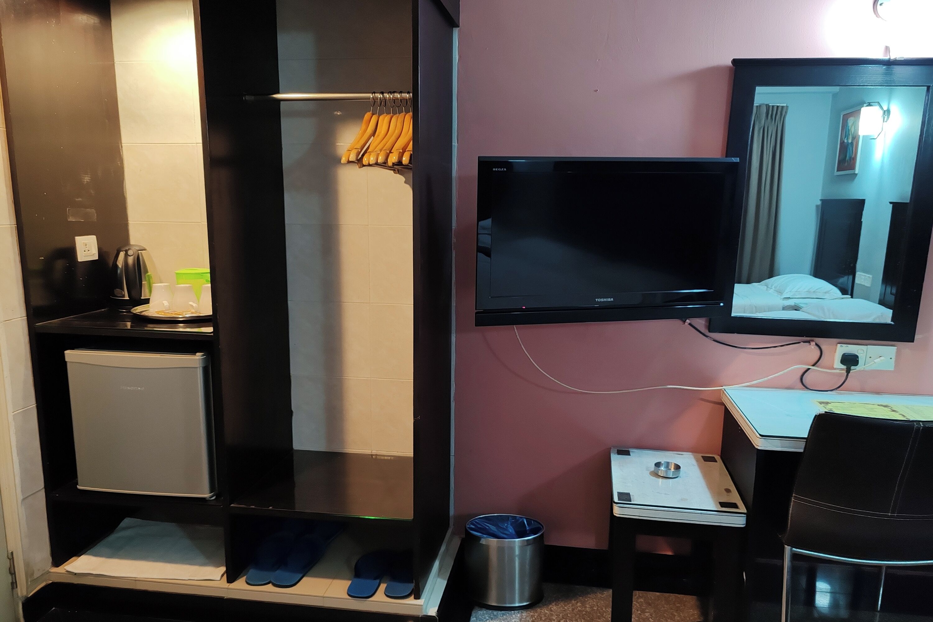 A hotel room with a shower, a small fridge, a TV, a desk with a chair, and a trash bin.